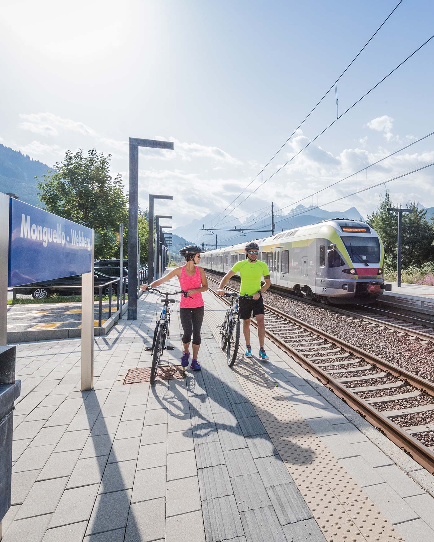 Zwei Radfahrer fahren mit ihren Fahrrädern an einem Bahnsteig entlang, während sich ein Zug unter einem hellen, sonnigen Himmel nähert. - Ansitz Toldt Dolomites Village