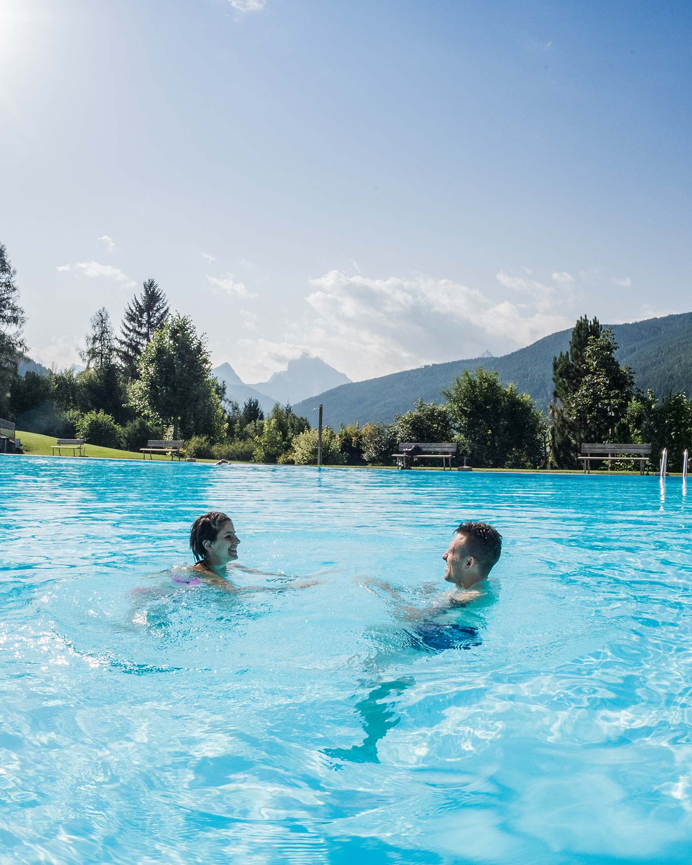Zwei Menschen schwimmen und lächeln in einem Pool mit Bergen und Bäumen im Hintergrund unter einem klaren Himmel. - Ansitz Toldt Dolomites Village