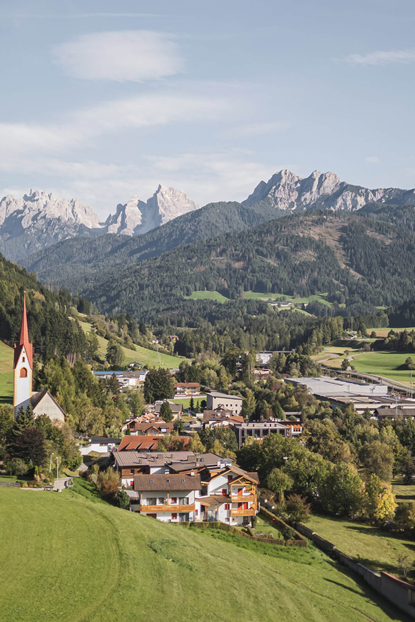 Ein Bergdorf mit einem Kirchturm, Häusern, grünen Feldern und bewaldeten Hügeln unter einem blauen Himmel. - Ansitz Toldt Dolomites Village