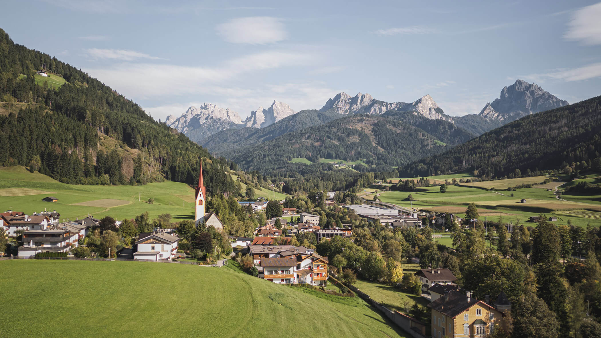 Bergdorf mit Kirchturm, Häusern, grünen Feldern und bewaldeten Hügeln unter blauem Himmel. - Ansitz Toldt Dolomites Village