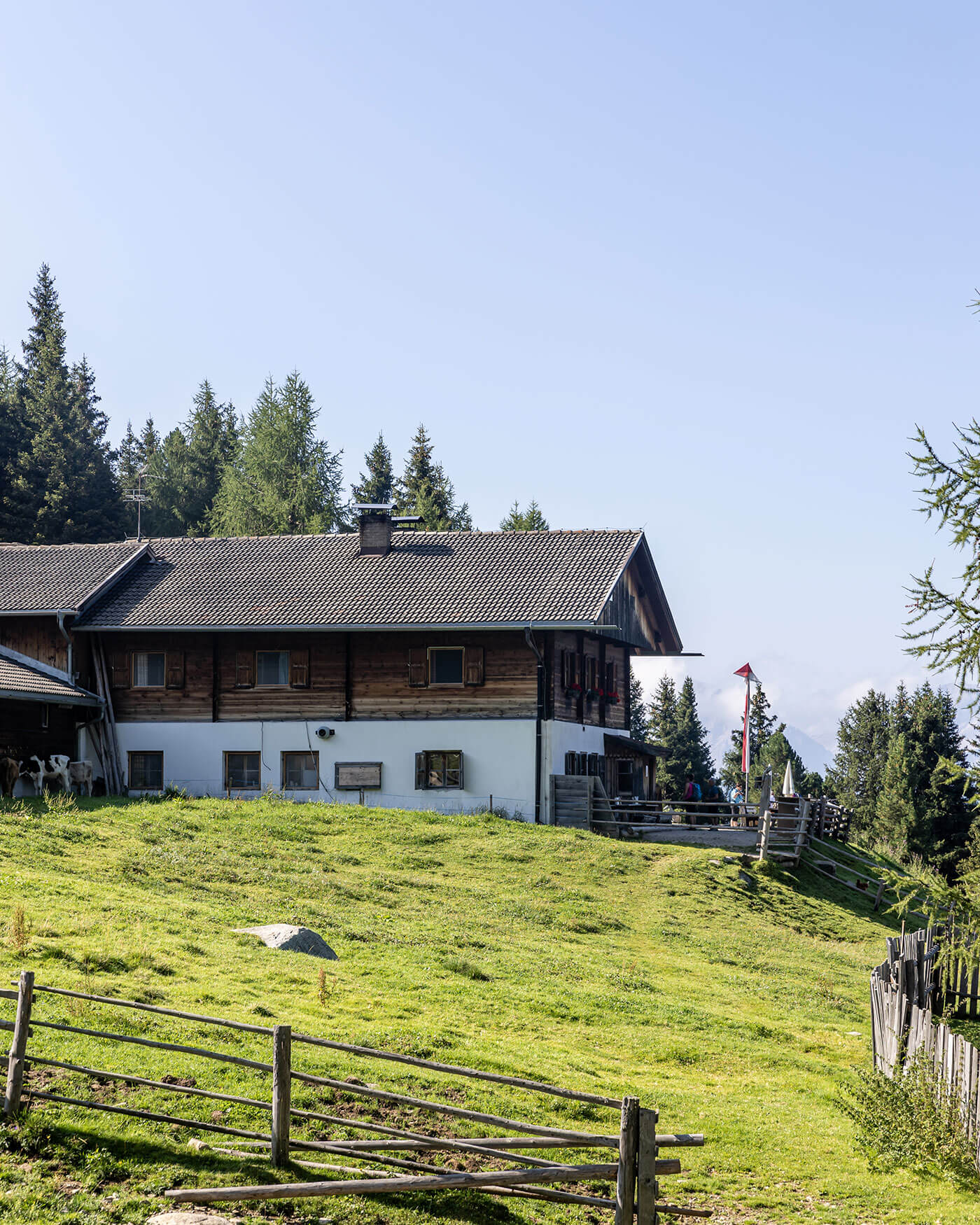 Ein rustikales Holzhaus steht auf einem grasbewachsenen Hügel mit einem Holzzaun und Bäumen im Hintergrund. - Ansitz Toldt Dolomites Village