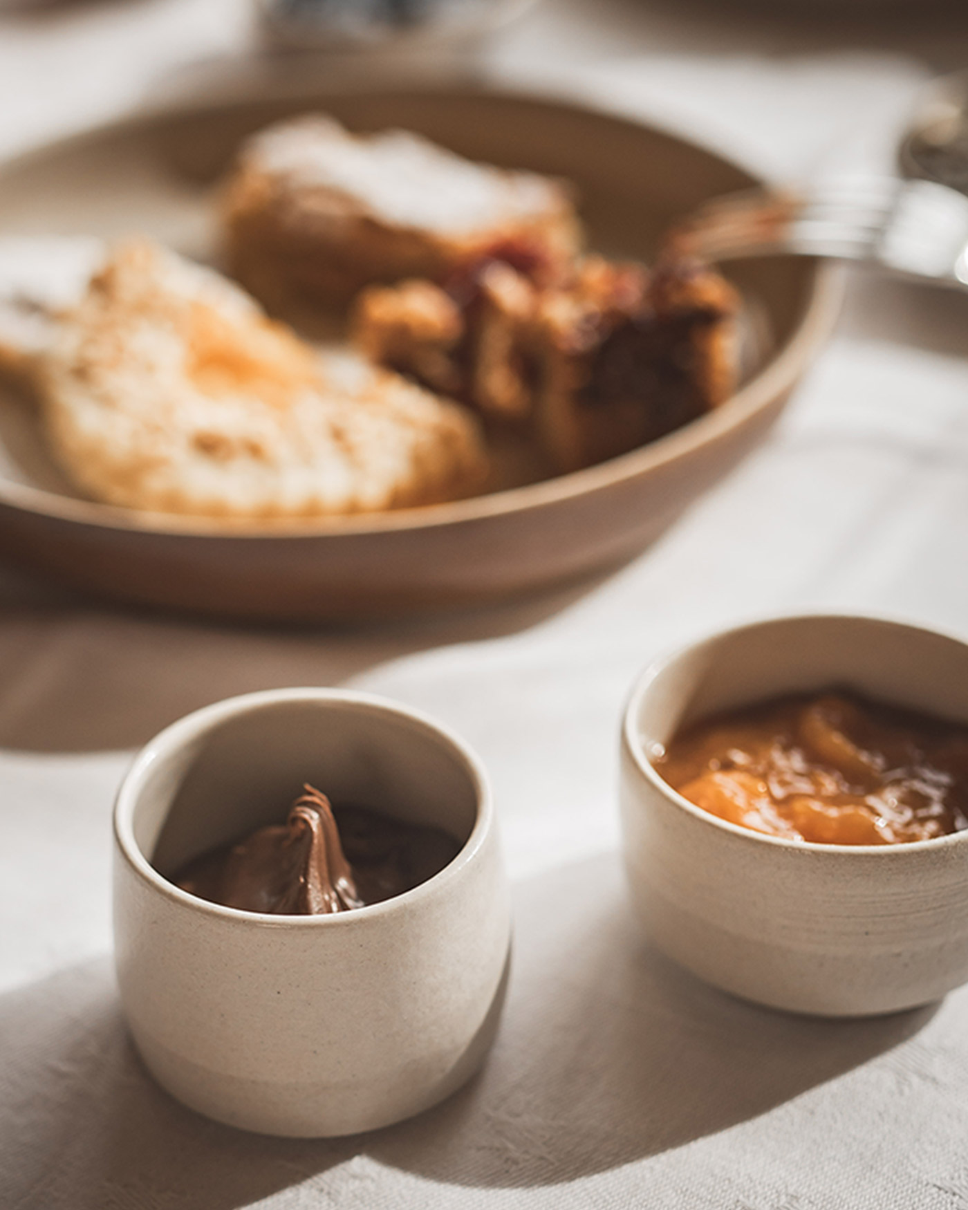 Zwei kleine Schälchen mit Schokoladenaufstrich und Orangenmarmelade, im Hintergrund ein Teller mit Gebäck. - Ansitz Toldt Dolomites Village