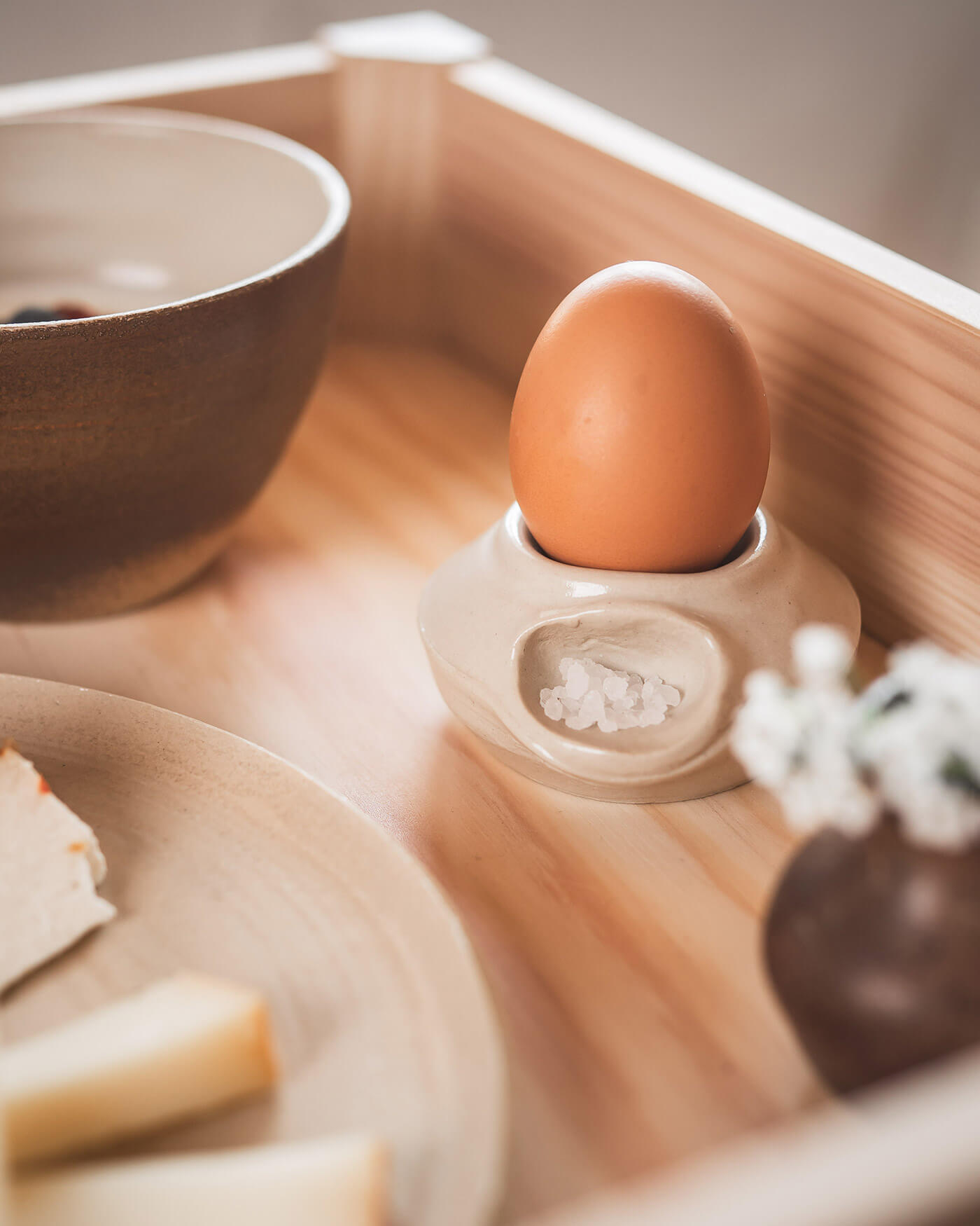A brown egg in a ceramic holder with salt, next to a bowl and plate on a wooden tray. - Ansitz Toldt Dolomites Village