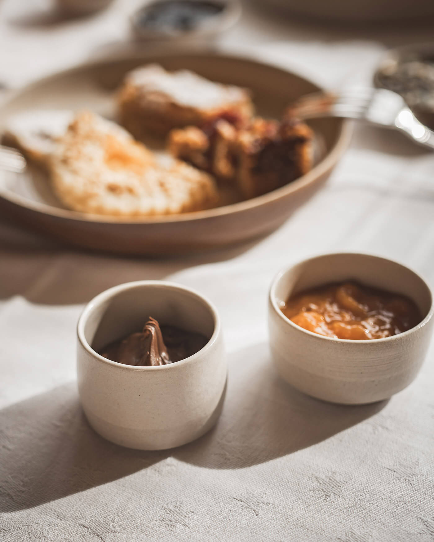 Two small bowls of chocolate spread and jam, with pastries on a plate in the background, on a sunlit table. - Ansitz Toldt Dolomites Village