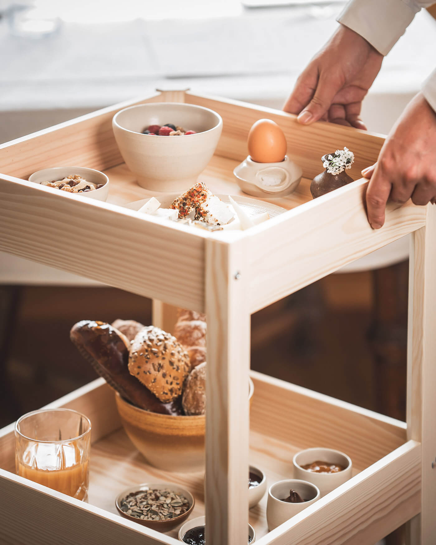 Eine Person stellt einen hölzernen Frühstückswagen mit Eiern, Joghurt, Brot, Nüssen und verschiedenen kleinen Speisen zusammen. - Ansitz Toldt Dolomites Village