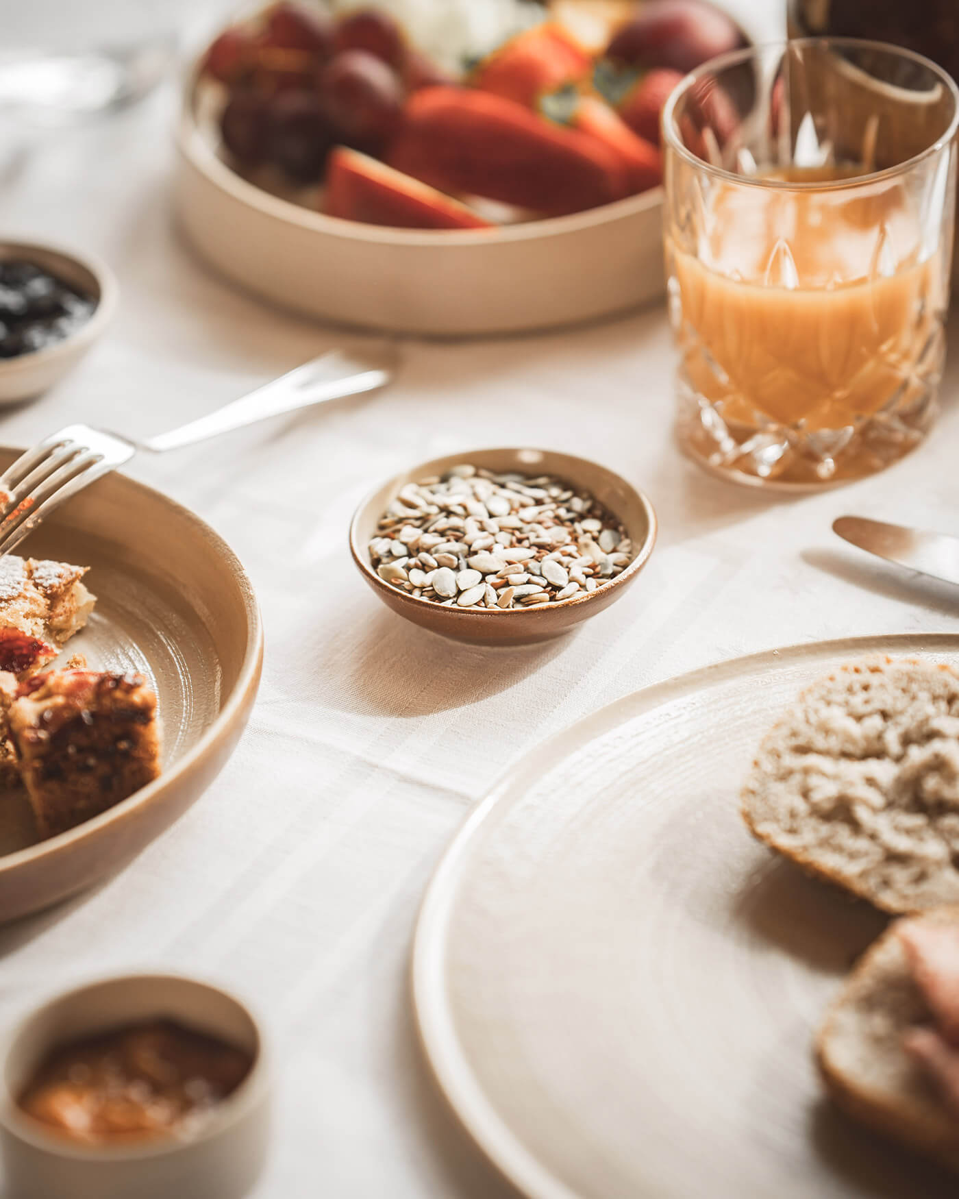 Frühstückstisch mit Gebäck, Saft, Obst, Samen und Marmelade auf neutralem Geschirr. - Ansitz Toldt Dolomites Village