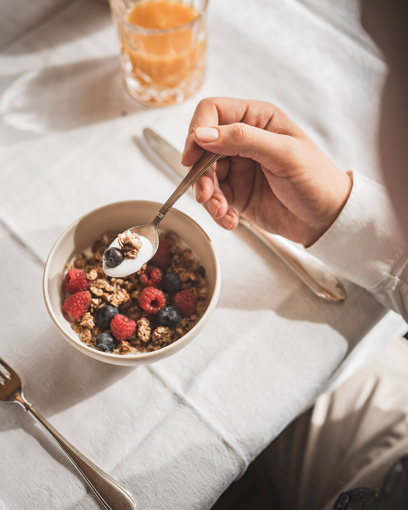 Eine Hand hält einen Löffel Müsli mit Joghurt und Beeren über einer Schüssel auf einem weißen Tischtuch. - Ansitz Toldt Dolomites Village