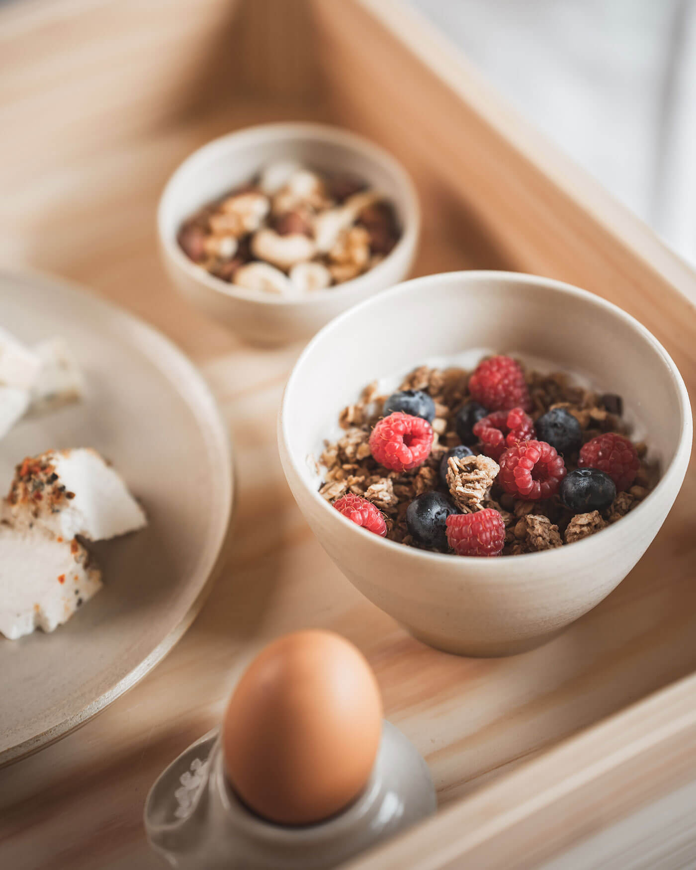 Eine Schale Müsli mit Himbeeren und Heidelbeeren, ein Ei und eine kleine Schale mit gemischten Nüssen auf einem Holztablett. - Ansitz Toldt Dolomites Village