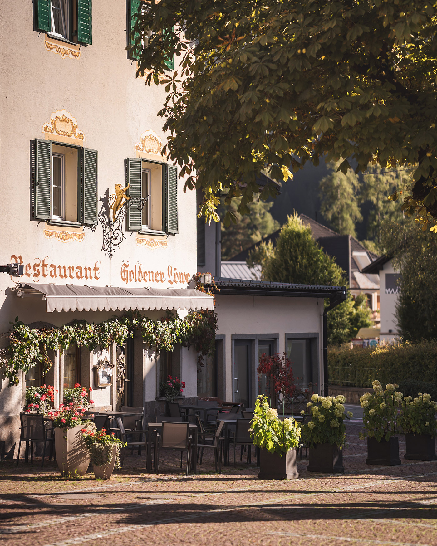 Ein charmantes Restaurant mit Sitzgelegenheiten im Freien und Blumen, im Schatten von Bäumen in einem sonnenbeschienenen europäischen Dorf. - Ansitz Toldt Dolomites Village