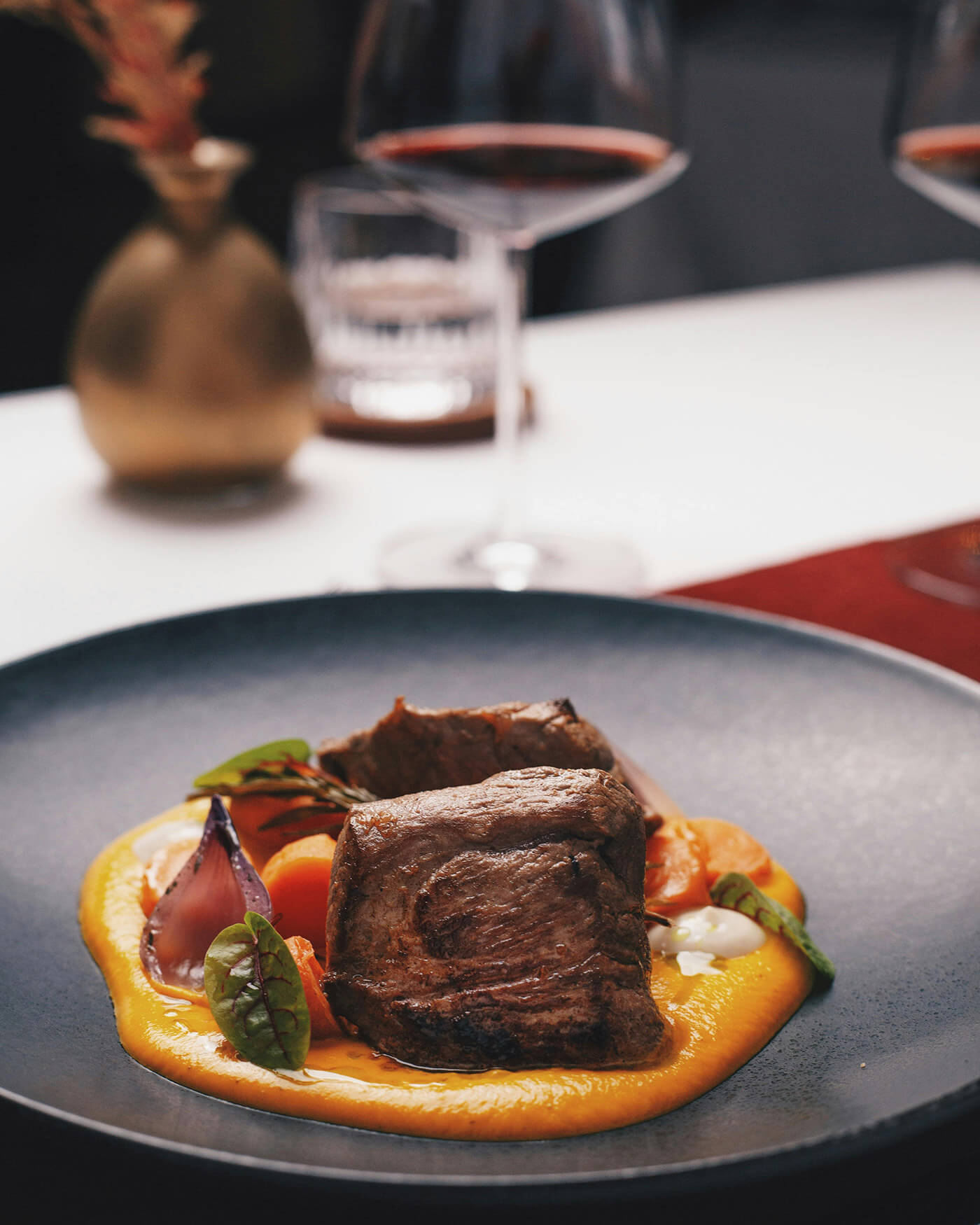 Ein Gourmet-Rindfleischgericht mit Gemüse und Soße, serviert auf einem dunklen Teller mit Weingläsern im Hintergrund. - Ansitz Toldt Dolomites Village