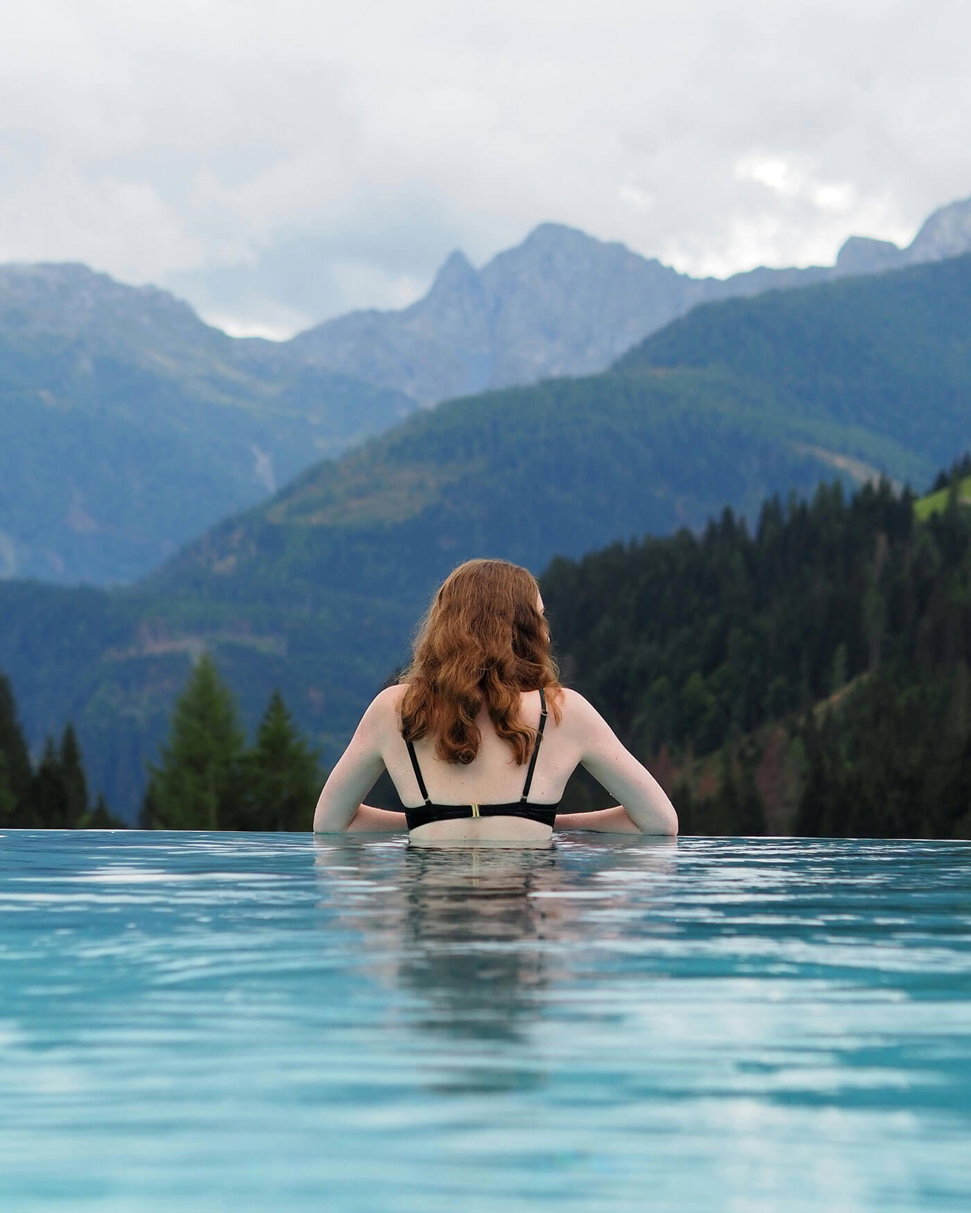Frau mit roten Haaren in einem Pool mit Blick auf bewaldete Berge und bewölkten Himmel im Hintergrund. - Ansitz Toldt Dolomites Village