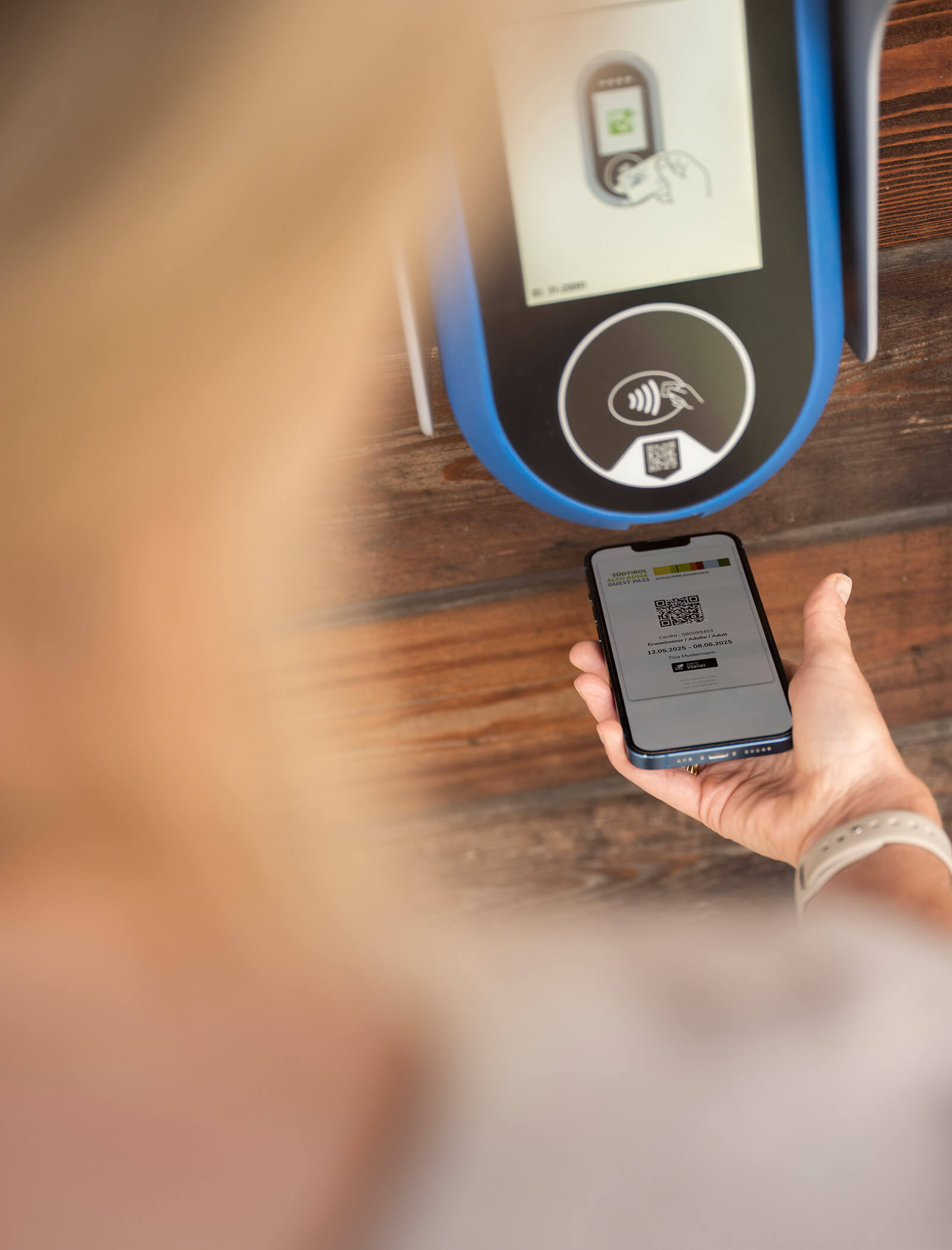 Eine Person hält ein Telefon mit einem QR-Code in der Nähe eines kontaktlosen Zahlungsterminals auf einer Holzoberfläche. - Ansitz Toldt Dolomites Village