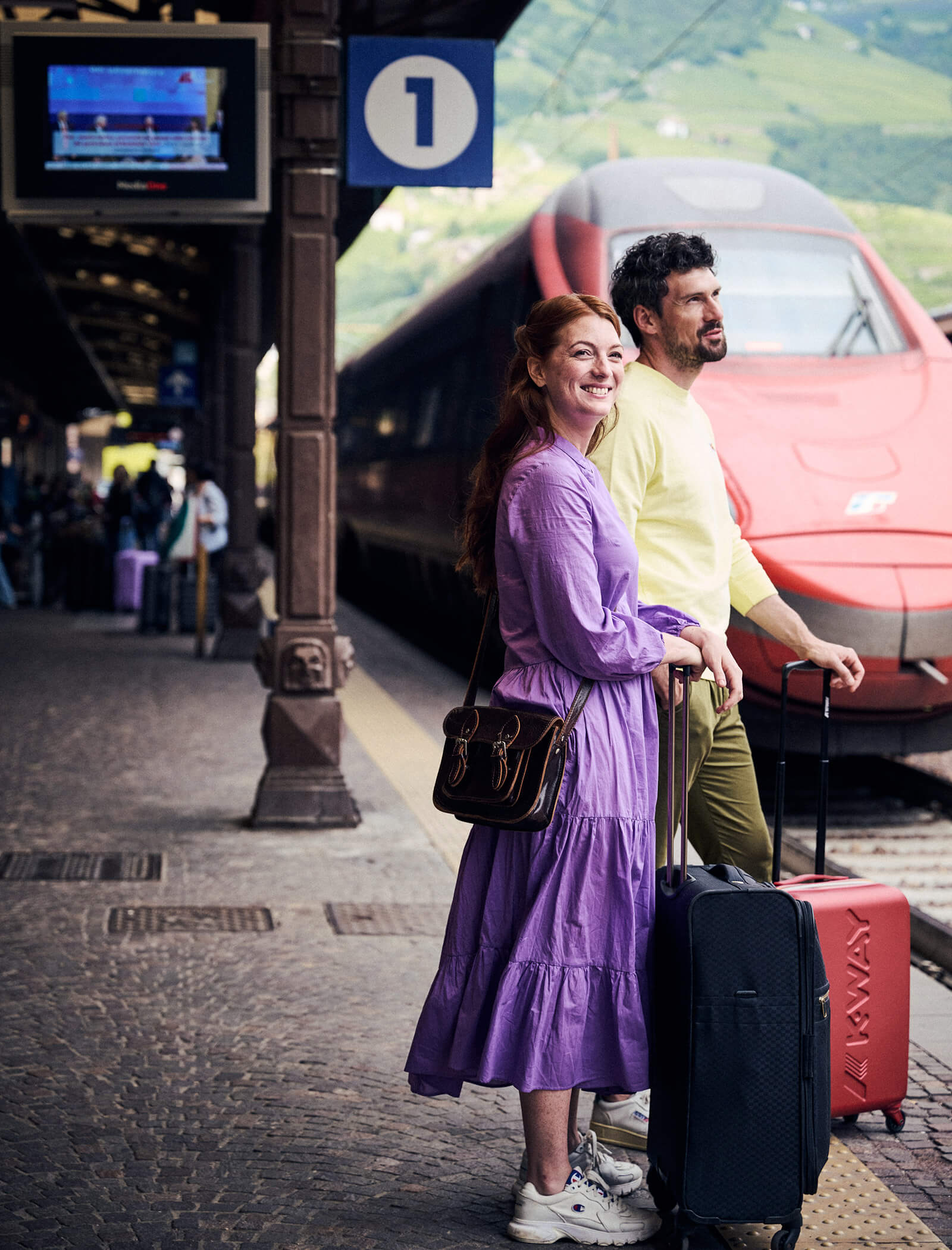 Zwei Personen mit Koffern stehen auf einem Bahnsteig neben einem roten Zug, lächeln und warten. - Ansitz Toldt Dolomites Village