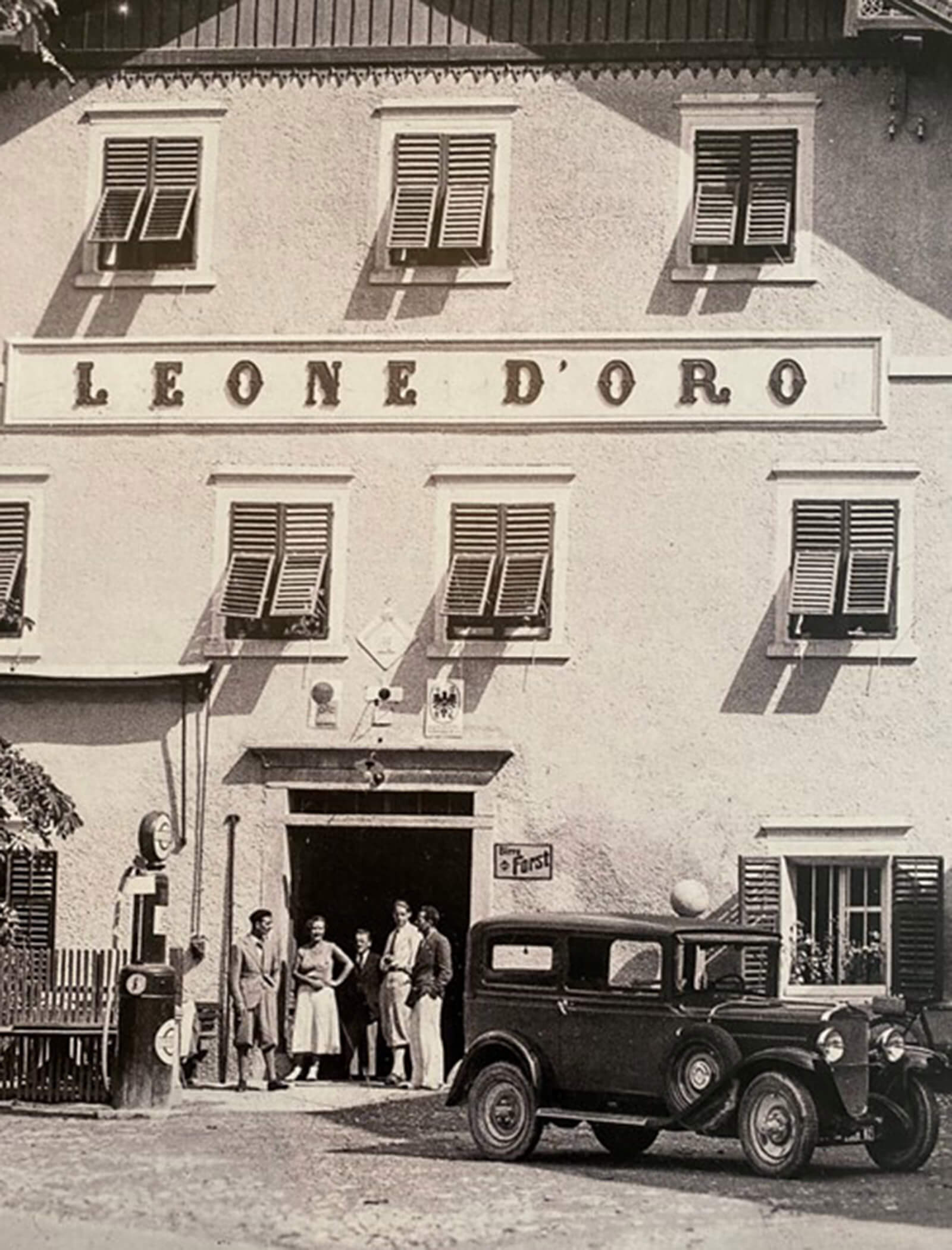 Schwarz-Weiß-Foto von Menschen und einem Oldtimer vor einem Gebäude mit der Aufschrift Leone DOro. - Ansitz Toldt Dolomites Village