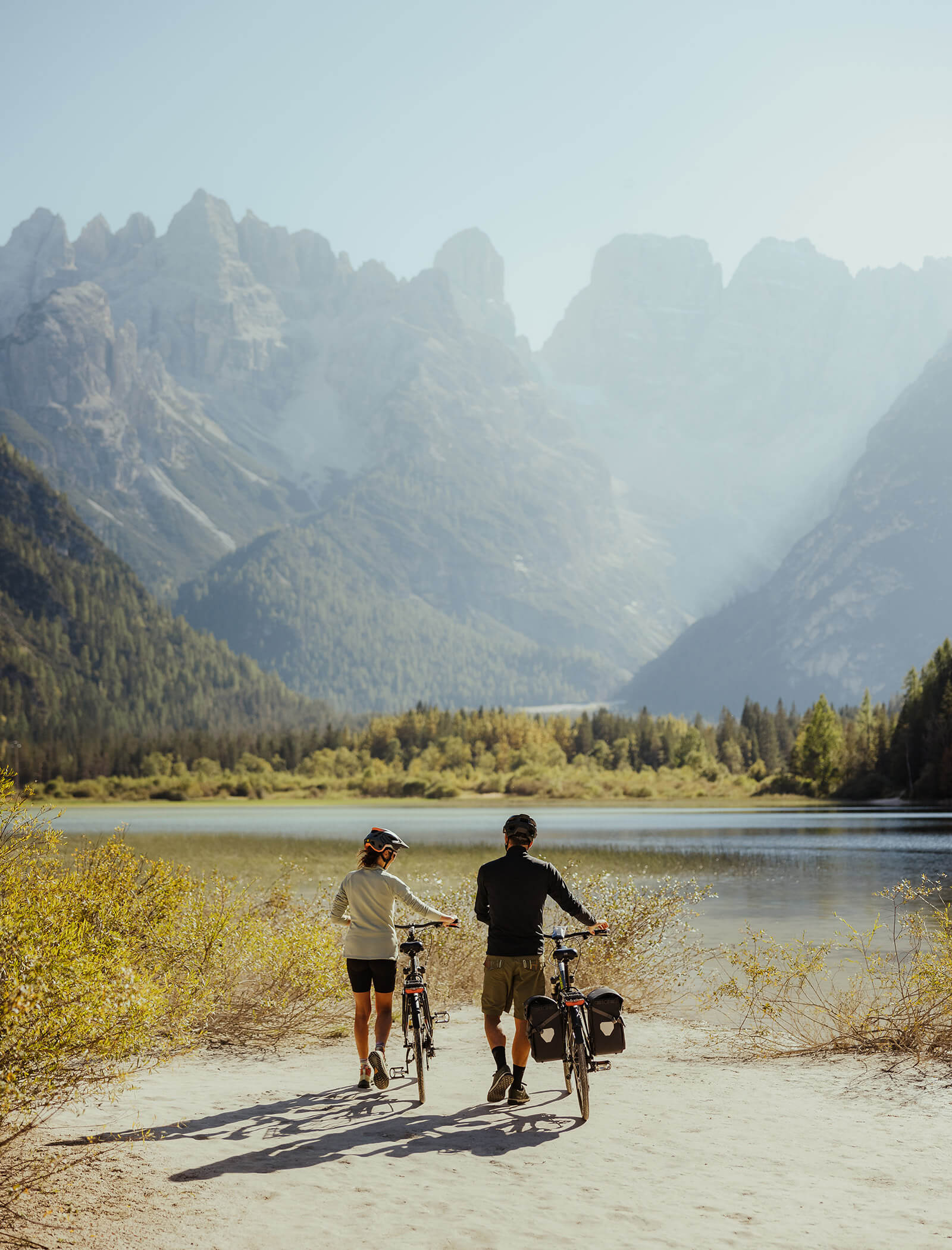 Zwei Radfahrer stehen an einem See mit Berggipfeln im Hintergrund, umgeben von Bäumen und Sonnenlicht. - Ansitz Toldt Dolomites Village