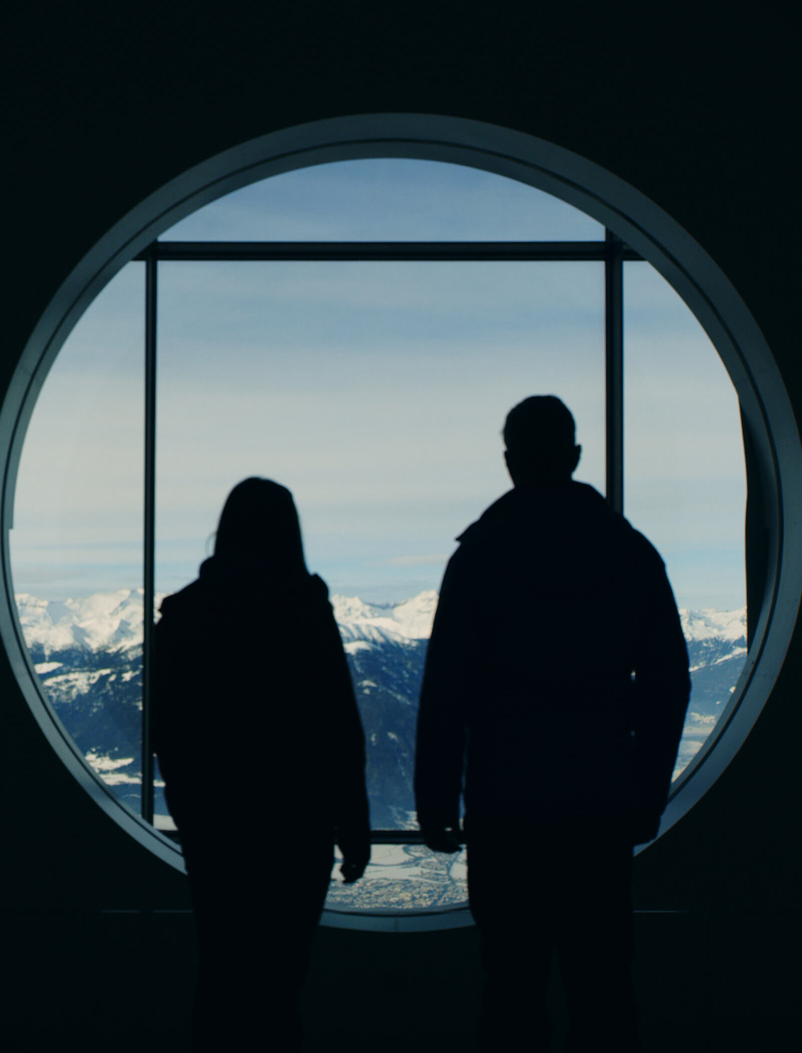 Die Silhouetten von zwei Personen stehen an einem runden Fenster mit Blick auf schneebedeckte Berge und einen blauen Himmel. - Ansitz Toldt Dolomites Village