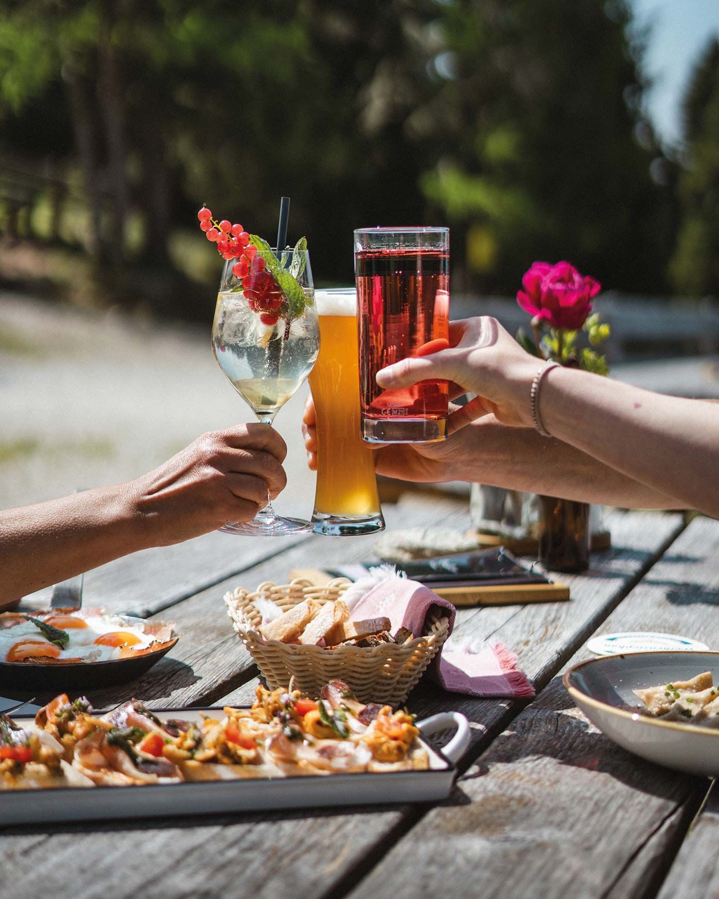 Drei Personen stoßen im Freien an einem Tisch mit Essen, Blumen und Sonnenlicht im Hintergrund auf Getränke an. - Ansitz Toldt Dolomites Village