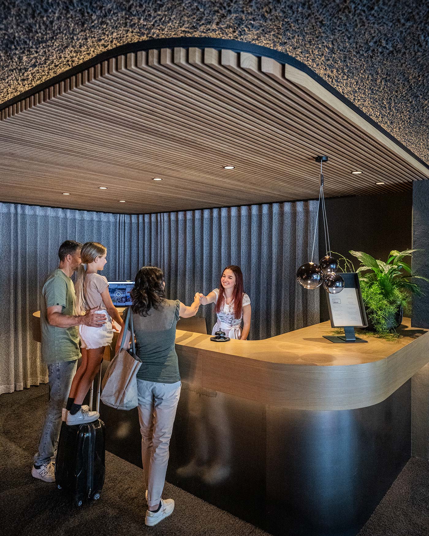 Eine Familie checkt an der Rezeption eines modernen Hotels mit lächelnder Rezeptionistin und modernem Dekor ein. - Ansitz Toldt Dolomites Village