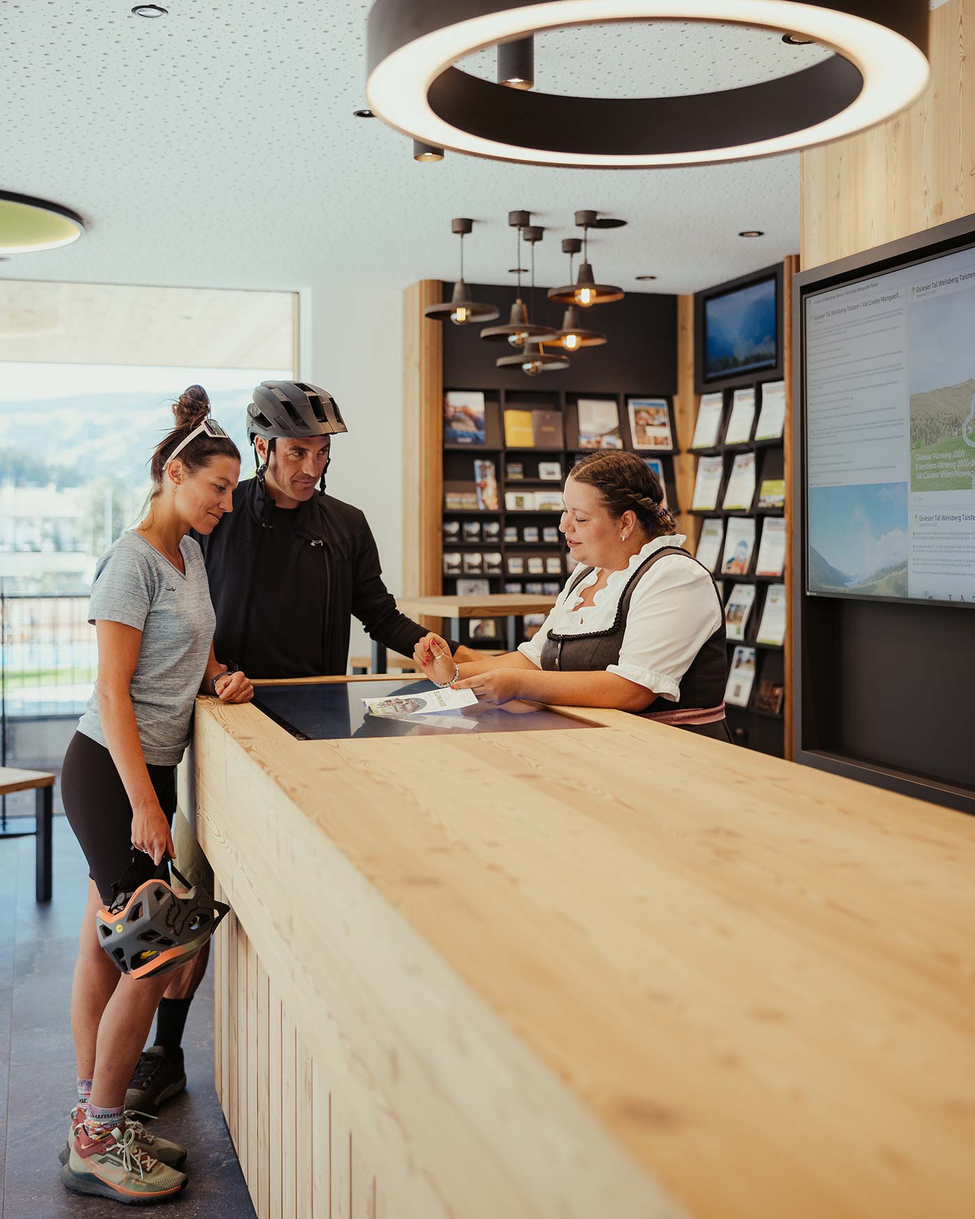 Zwei Radfahrer mit Helmen unterhalten sich mit einem Rezeptionisten an einem hölzernen Schalter in einem modernen Informationszentrum. - Ansitz Toldt Dolomites Village
