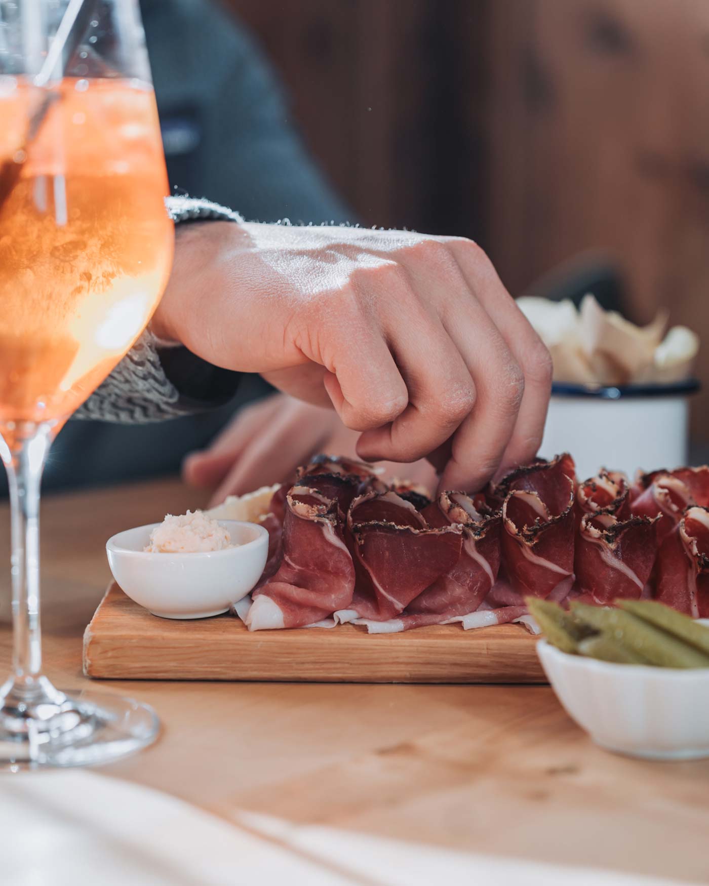 Die Hand greift nach gepökeltem Fleisch auf einem Charcuterie-Brett mit Essiggurken, Dip und einem Glas Orangengetränk in der Nähe. - Ansitz Toldt Dolomites Village