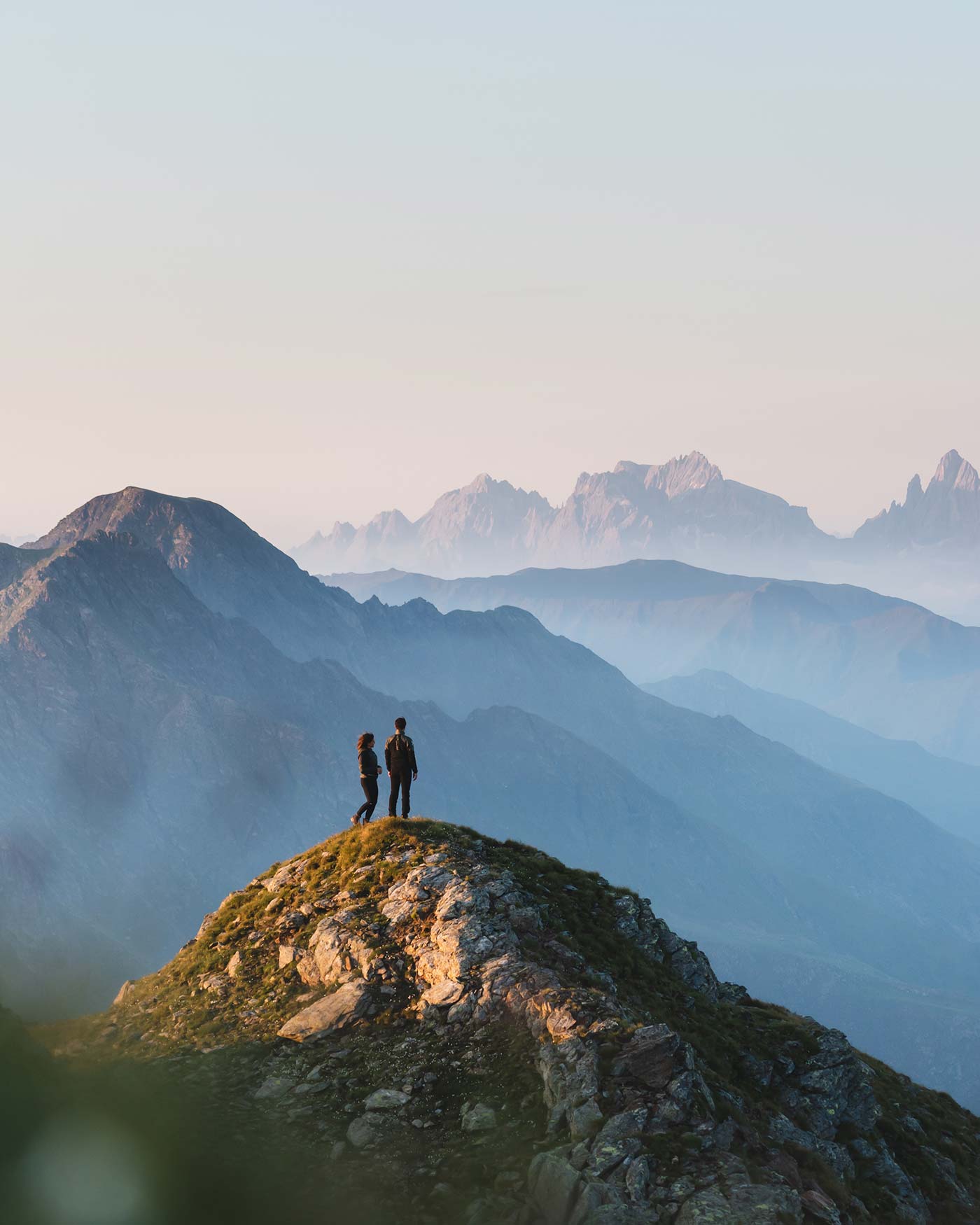 Zwei Personen stehen auf einem Berggipfel und überblicken bei Sonnenaufgang oder Sonnenuntergang die nebelverhangene Berglandschaft. - Ansitz Toldt Dolomites Village