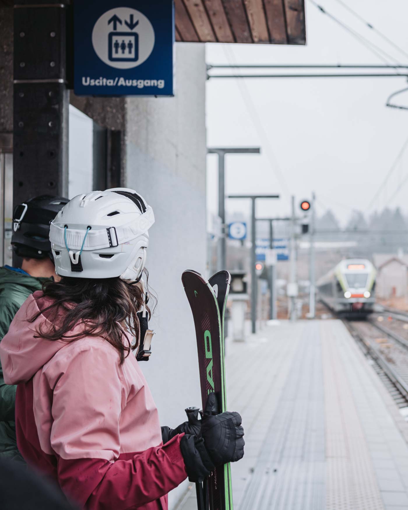 Menschen in Skiausrüstung warten am Bahnsteig eines Bahnhofs, während sich in der Ferne ein Zug nähert. - Ansitz Toldt Dolomites Village