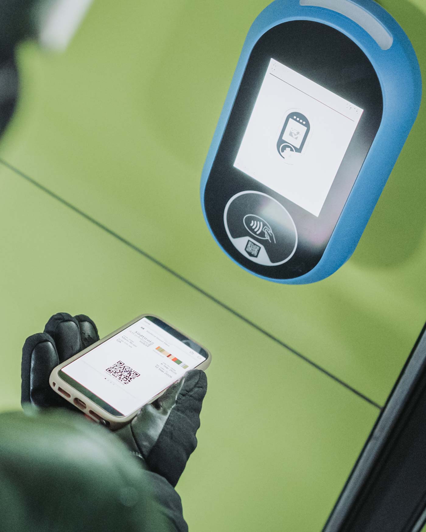 Eine behandschuhte Hand hält ein Telefon mit einem QR-Code in der Nähe eines kontaktlosen Zahlungsterminals auf grünem Hintergrund. - Ansitz Toldt Dolomites Village