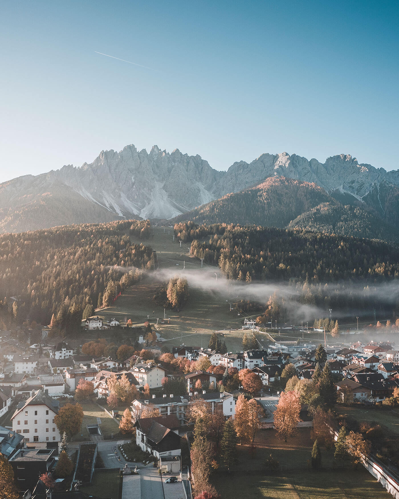 Ein Bergdorf mit herbstlichen Bäumen und Nebel, unter zerklüfteten Gipfeln und einem klaren blauen Himmel. - Ansitz Toldt Dolomites Village