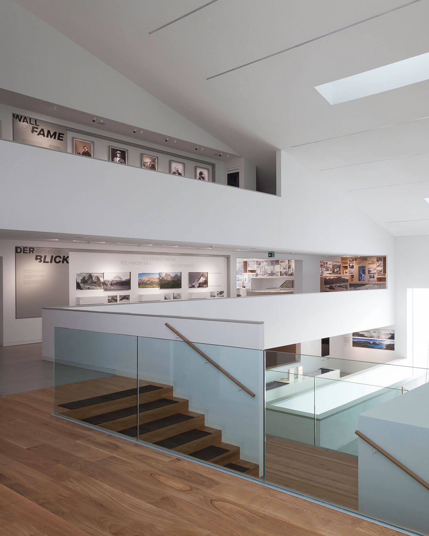 Moderne Kunstgalerie mit weißen Wänden, Holztreppe, Glasgeländer und Fotoauslagen auf zwei Ebenen. - Ansitz Toldt Dolomites Village