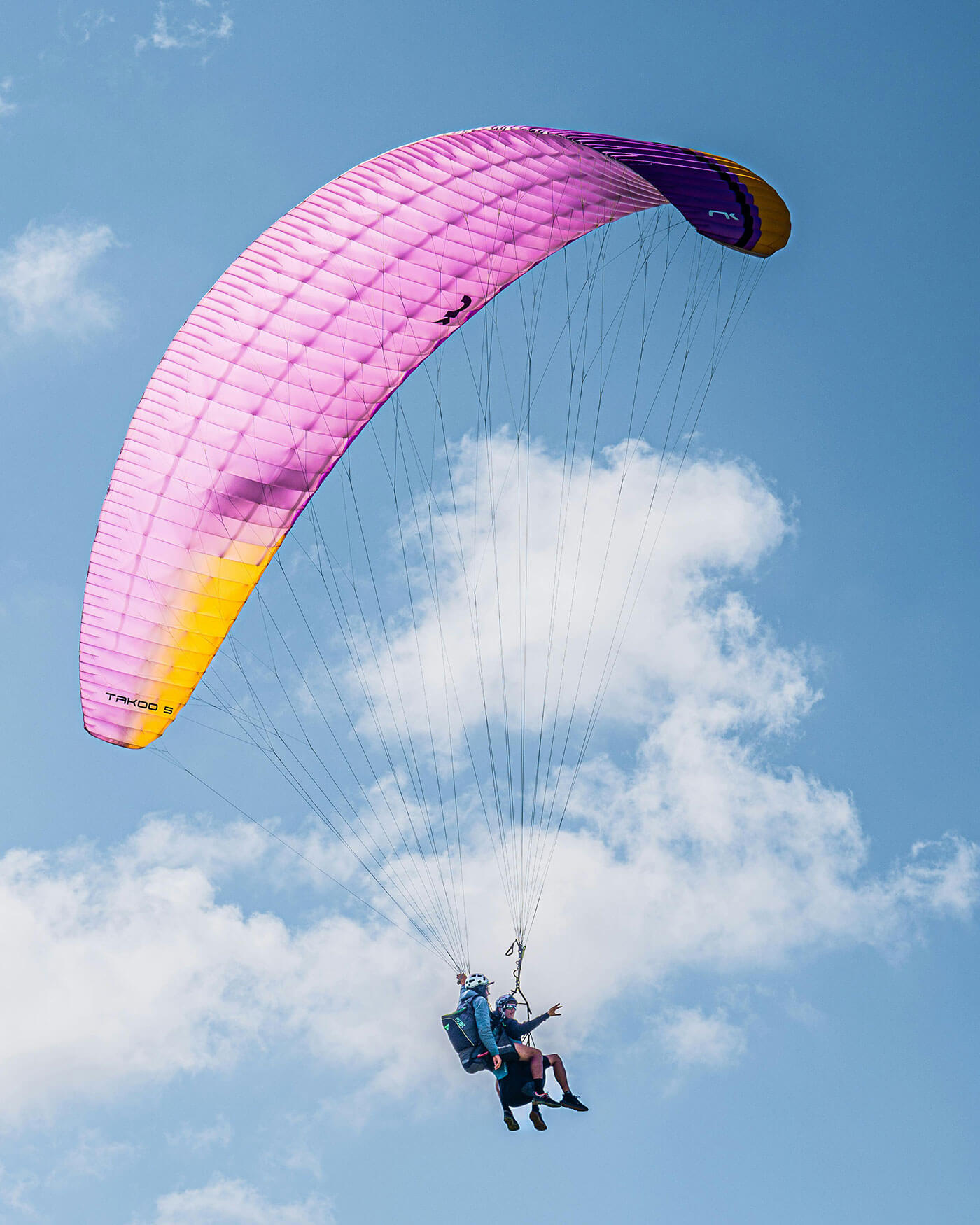 Zwei Personen beim Gleitschirmfliegen unter einem rosa-gelben Fallschirm vor einem blauen Himmel mit vereinzelten Wolken. - Ansitz Toldt Dolomites Village