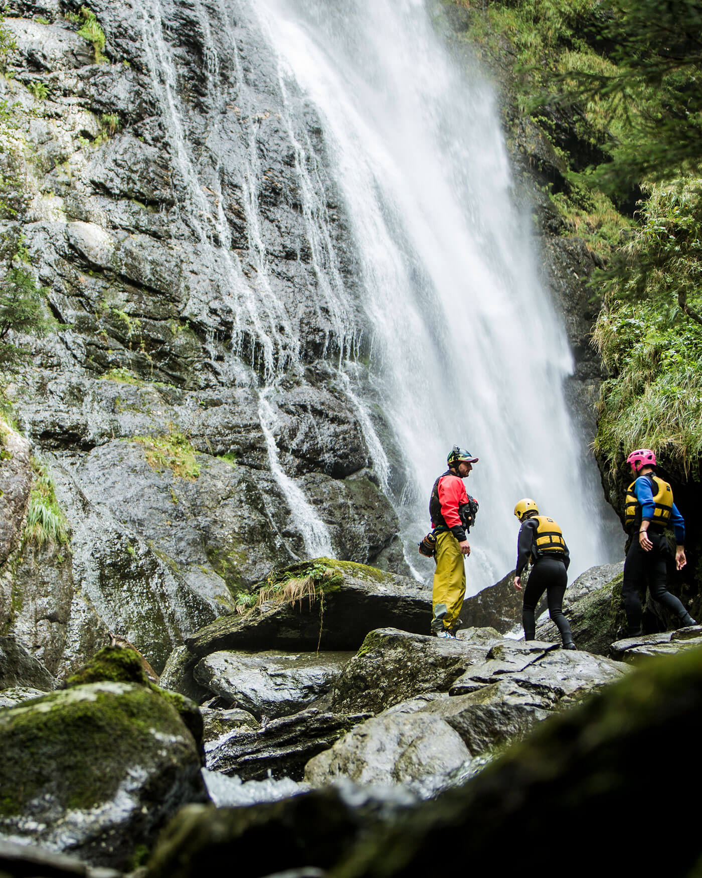 Drei Personen mit Helmen und Schwimmwesten stehen auf Felsen an einem großen Wasserfall in einem Wald. - Ansitz Toldt Dolomites Village