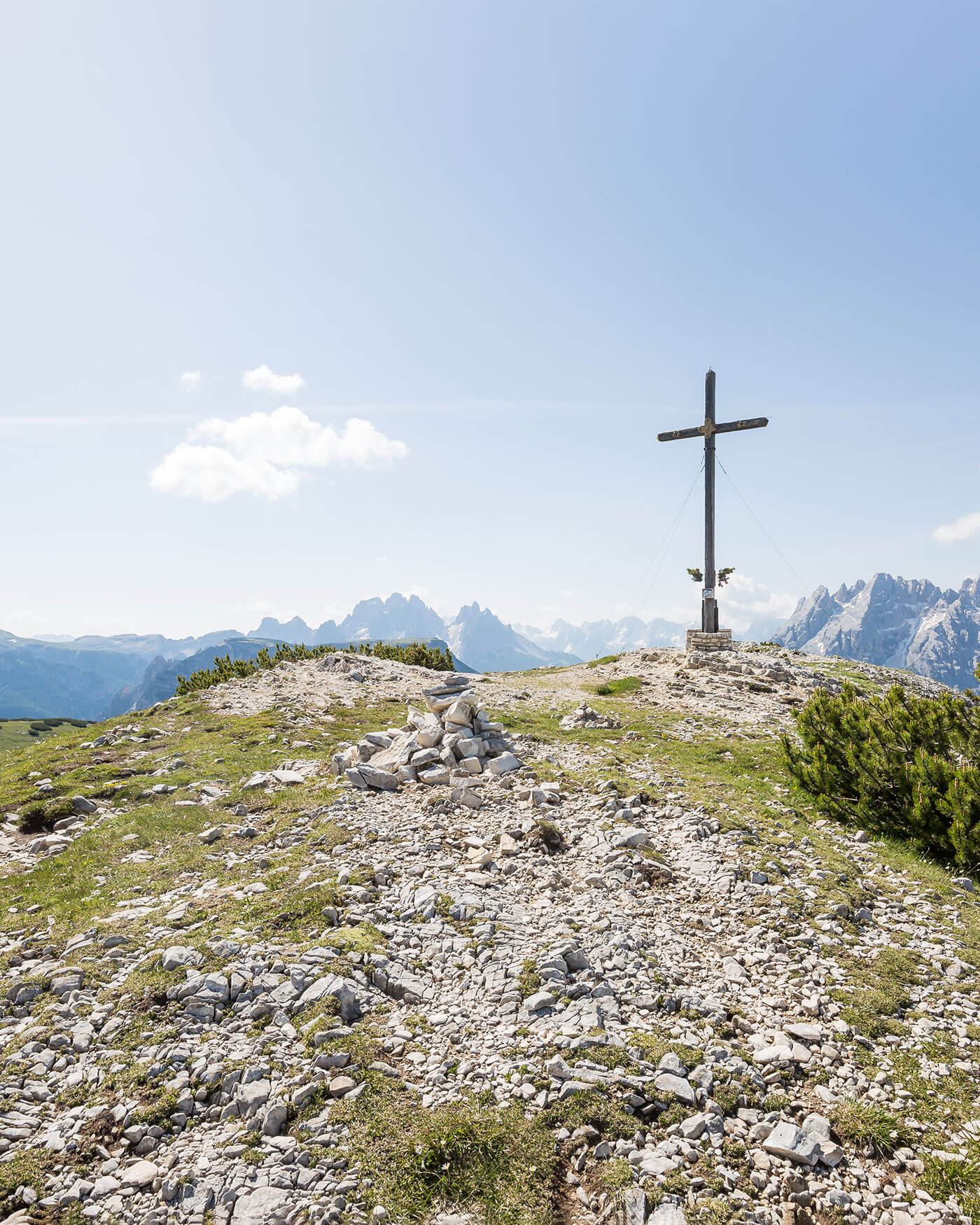 Ein Holzkreuz steht auf einer felsigen Bergkuppe mit Bergen und einem klaren Himmel im Hintergrund. - Ansitz Toldt Dolomites Village