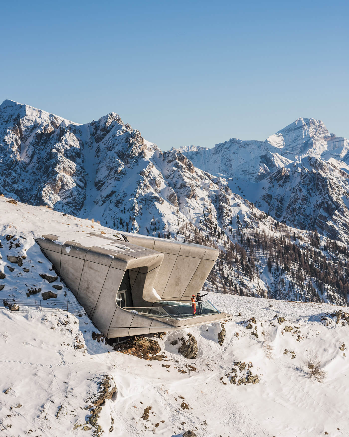 Ein modernes Betongebäude ragt aus einem verschneiten Berghang, auf dessen gläsernem Balkon eine Person steht. - Ansitz Toldt Dolomites Village