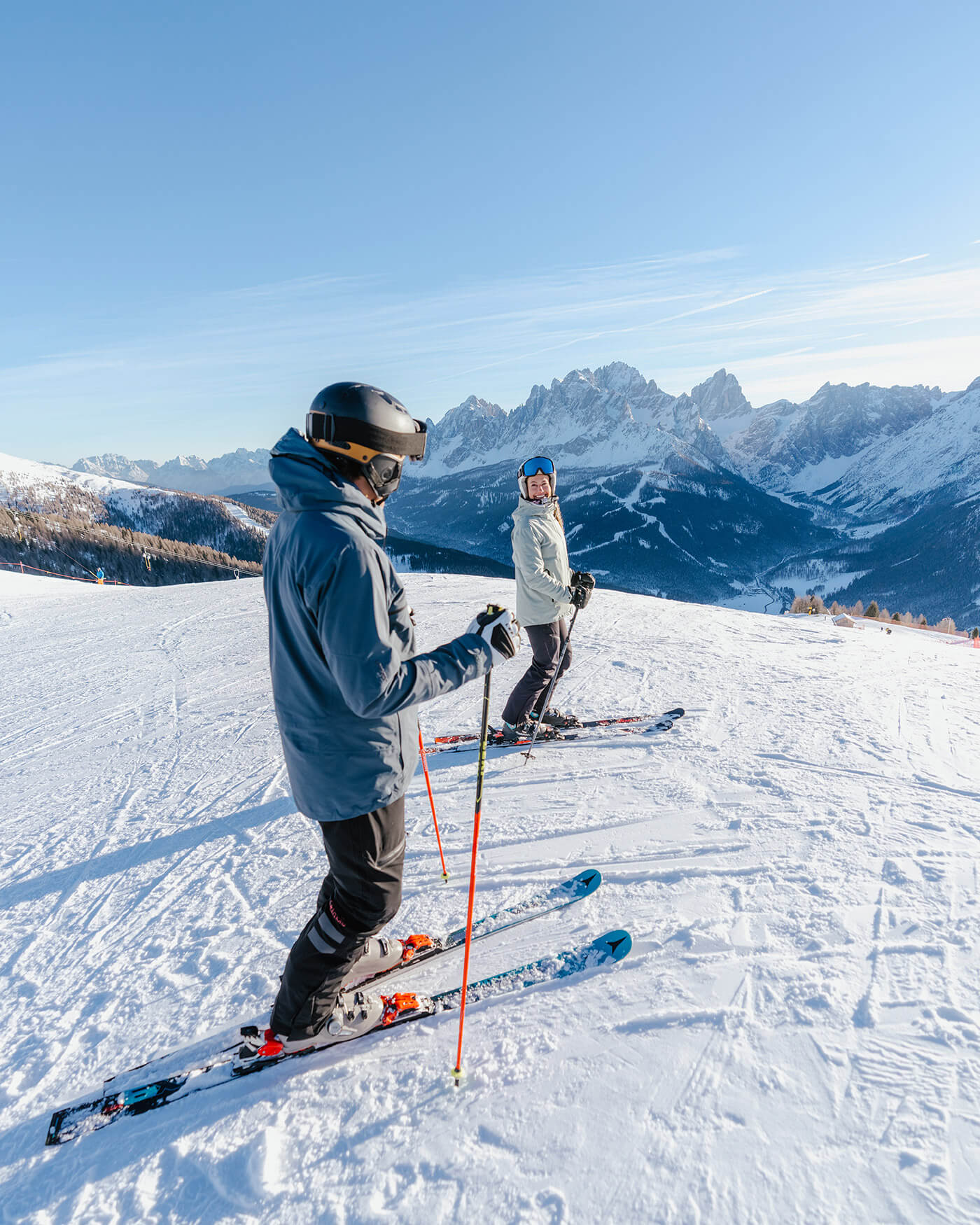 Zwei Personen beim Skifahren auf einem verschneiten Berg mit malerischen, sonnenbeschienenen Gipfeln im Hintergrund. - Ansitz Toldt Dolomites Village