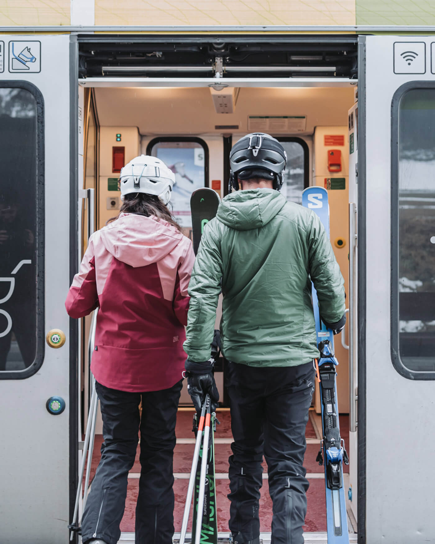 Zwei Personen in Skikleidung und mit Helmen besteigen einen Zug und tragen Skier und Stöcke, von hinten gesehen. - Ansitz Toldt Dolomites Village